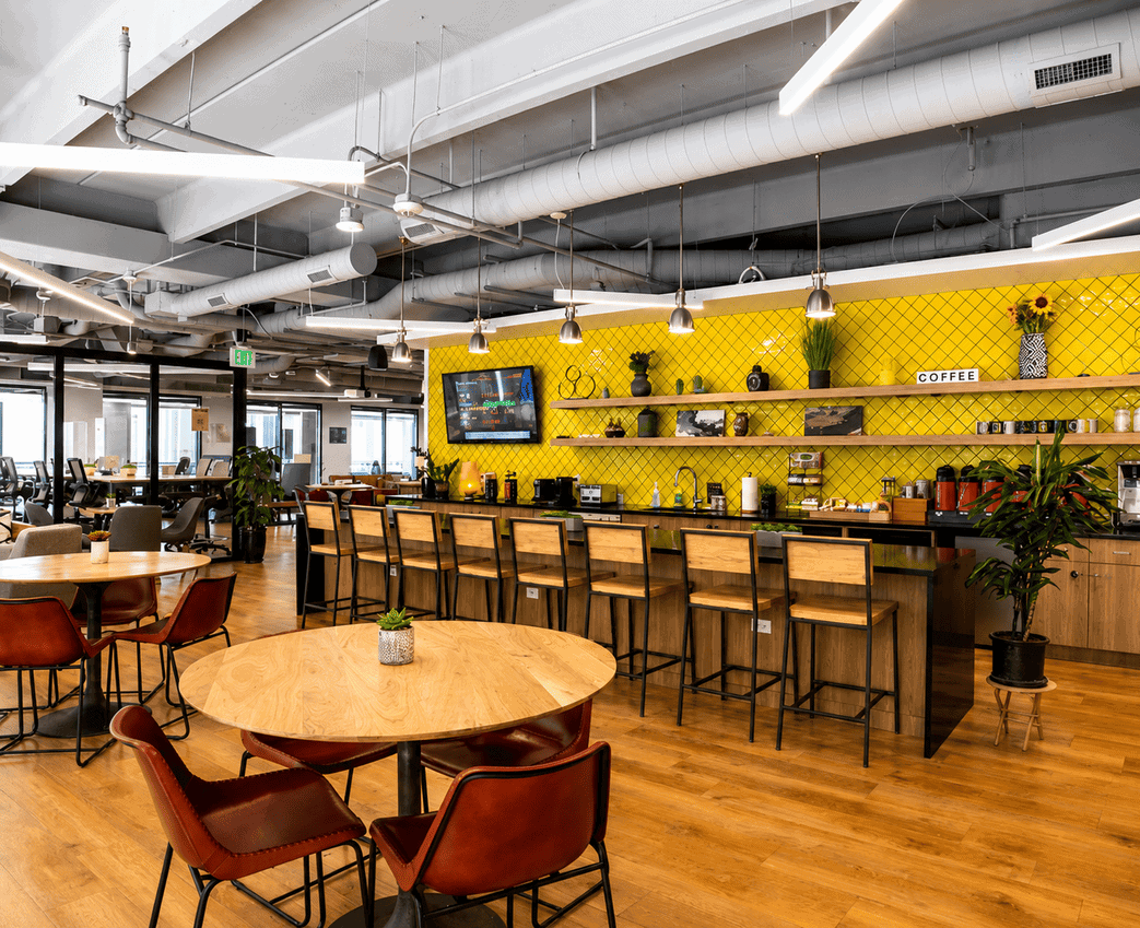 Modern coworking space with a vibrant yellow accent wall, bar seating, and communal tables, ideal for collaboration and networking.
