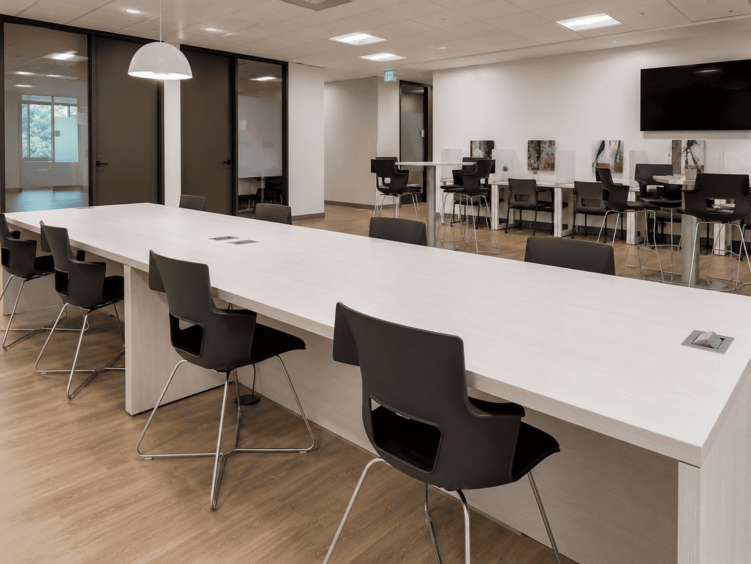 Modern coworking space with a long white table, black chairs, and private meeting rooms with glass walls.
